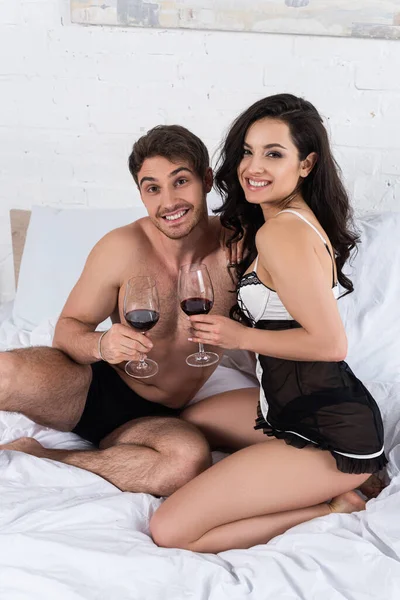Mujer y hombre con copas de vino tinto mirando a la cámara y sonriendo en la cama en el dormitorio - foto de stock
