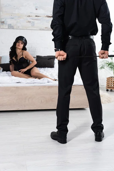 Attractive policewoman in peaked cap on bed looking at man with flogging whip in bedroom — Stock Photo