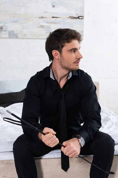 Man with flogging whip looking away and sitting on bed — Stock Photo