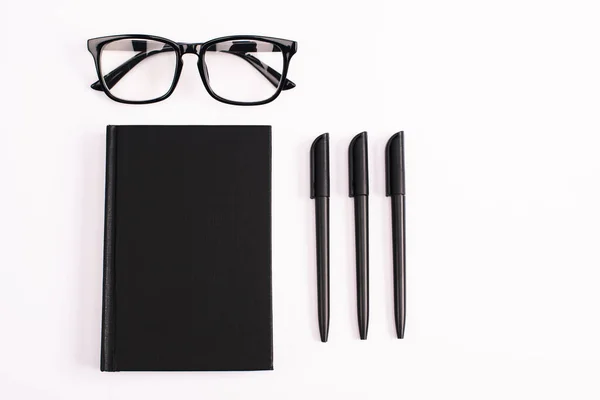 Couché plat avec des stylos près des lunettes et carnet noir isolé sur blanc — Photo de stock