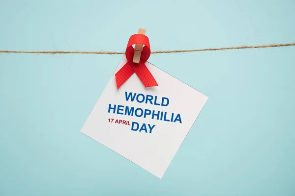 Card with world hemophilia day lettering and red ribbon on cord with pin isolated on blue — Stock Photo