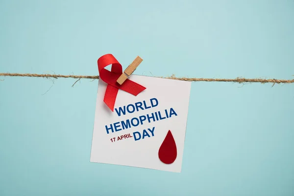 Card with world hemophilia day lettering and red ribbon on rope with pin isolated on blue — Stock Photo