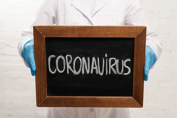 Обрезанный вид доктора, держащего доску с коронавирусной надписью на белом фоне — стоковое фото