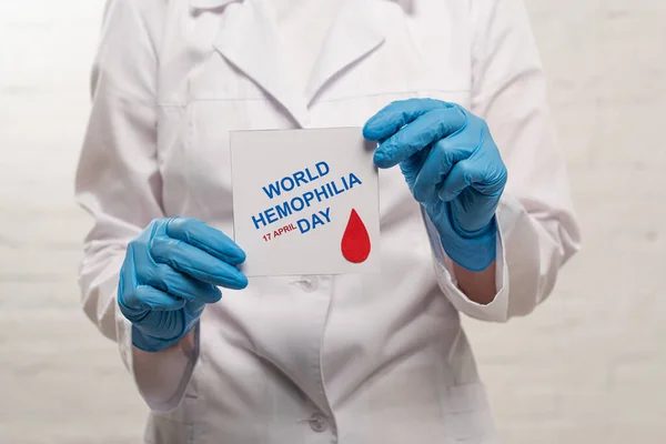 Vista recortada del médico en guantes de látex que sostiene la tarjeta con el día mundial de la hemofilia sobre fondo blanco - foto de stock