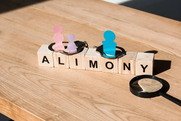 Wooden blocks with alimony lettering near family paper cut, magnifier and handcuffs on surface — Stock Photo