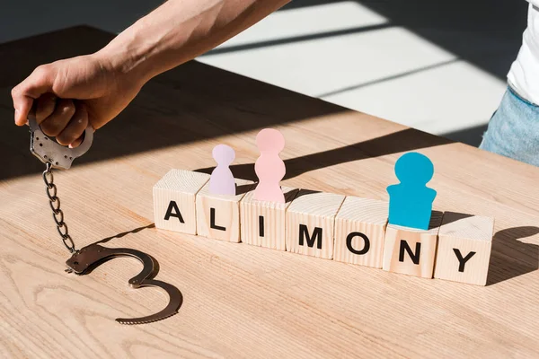 Cropped view of man holding handcuffs near wooden blocks with alimony lettering and paper people on desk — Stock Photo