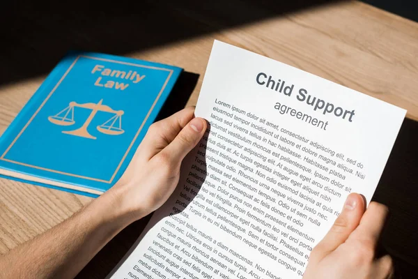 Vista ritagliata di uomo in possesso di documento con accordo di mantenimento dei figli vicino al libro di diritto di famiglia sulla scrivania — Foto stock