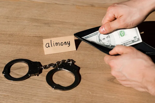 Обрезанный вид человека, держащего бумажник и долларовую банкноту возле бумаги с надписью алиментов и наручниками — стоковое фото