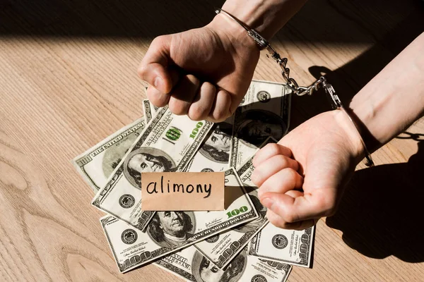 Обрезанный вид человека в наручниках рядом с бумагой с надписью алиментов и банкнотами в долларах — стоковое фото