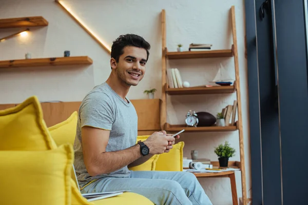 Enfoque selectivo del hombre con teléfono inteligente sonriendo y mirando a la cámara en la sala de estar - foto de stock