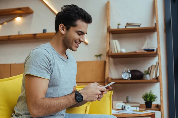 Mann plaudert auf Smartphone und lächelt auf Sofa im Wohnzimmer — Stockfoto