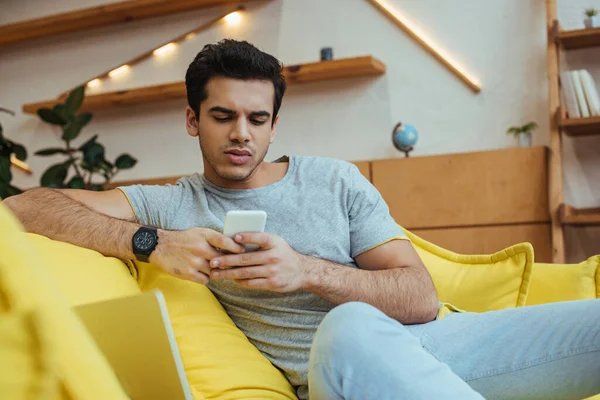 Selective focus of concentrated man chatting on smartphone on sofa in living room — Stock Photo