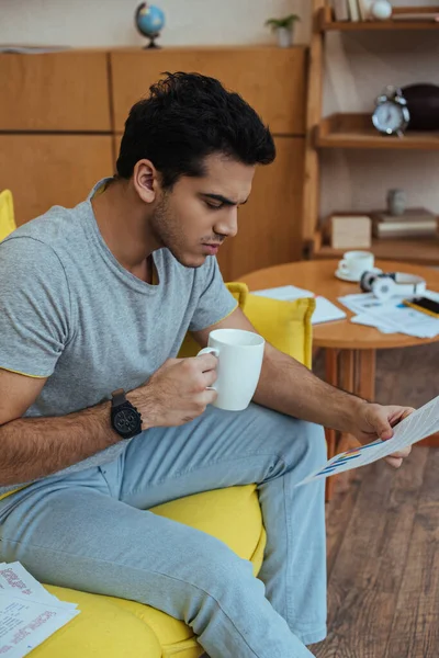 Freelancer with cup of tea looking at paper and sitting on sofa in living room — Stock Photo