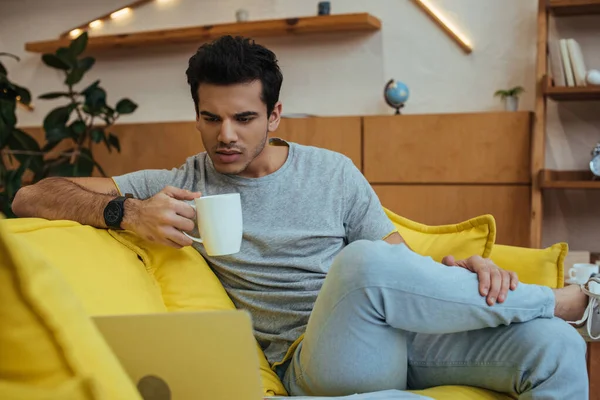 Selective focus of freelancer with cup of tea looking at laptop on sofa in living room — Stock Photo