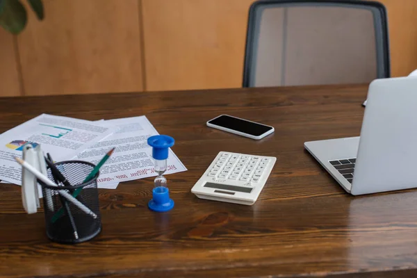 Papers, pen holder, hourglass, calculator and smartphone near laptop on table in office — Stock Photo