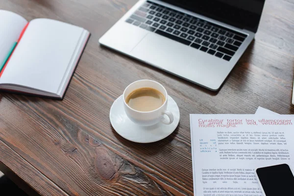 Vista de ángulo alto de la taza de café cerca de copybook, ordenador portátil y teléfono inteligente en los papeles en la mesa en la oficina - foto de stock