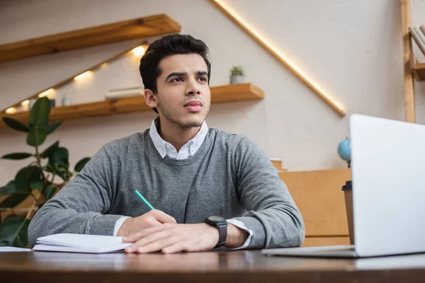 Low angle view of thoughtful businessman looking away and writing in notebook near laptop at table — Stock Photo