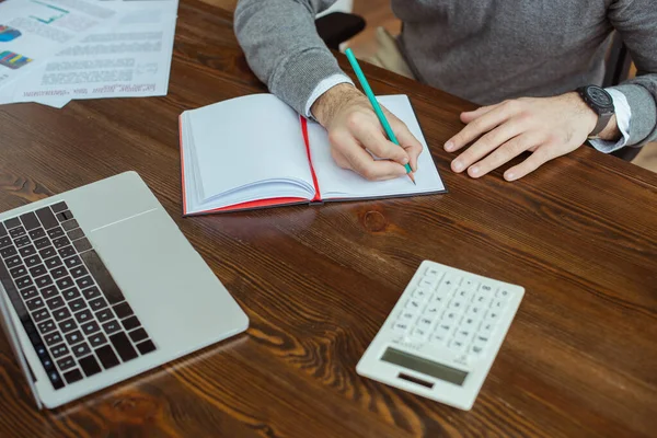 Vista cortada de homem de negócios escrevendo em notebook perto de laptop, calculadora e papéis à mesa no escritório — Fotografia de Stock