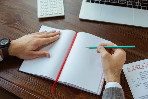 Vista parcial del hombre de negocios escribiendo en un cuaderno cerca de la computadora portátil y la calculadora en la mesa en la oficina - foto de stock