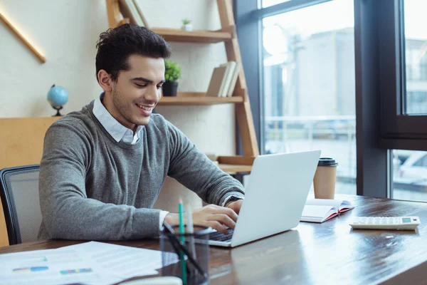 Избранное внимание бизнесмена, улыбающегося и работающего с ноутбуком за столом в офисе — стоковое фото