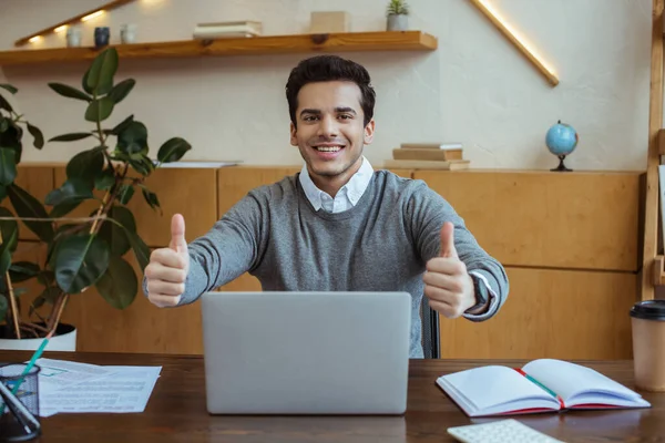 Бизнесмен с большими пальцами вверх улыбается и смотрит в камеру возле ноутбука за столом в офисе — стоковое фото