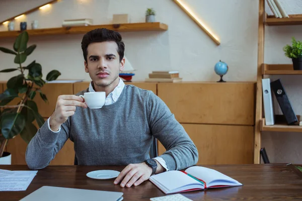 Бизнесмен держит чашку кофе и смотрит в камеру за столом в офисе — стоковое фото