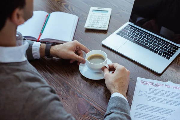 Vista recortada del hombre de negocios con taza de café cerca de la computadora portátil, calculadora, papeles y cuaderno en la mesa - foto de stock