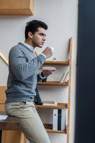 Selective focus of thoughtful businessman with saucer and cup of coffee in office — Stock Photo