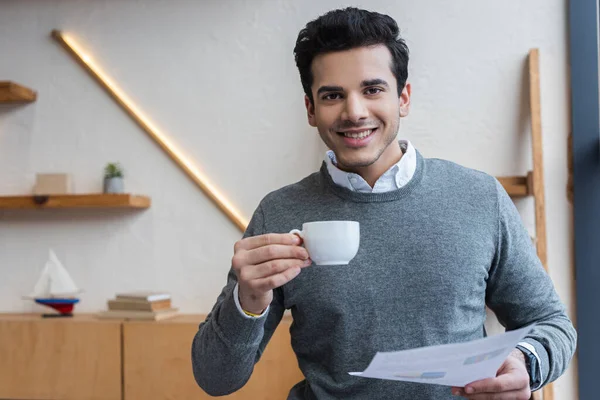Uomo d'affari con tazza di caffè e carta sorridente e guardando la macchina fotografica in ufficio — Foto stock