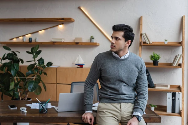 Бизнесмен смотрит в сторону и сидит на столе рядом с ноутбуком в офисе — стоковое фото