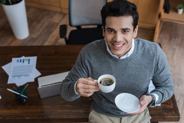 Vista de alto ángulo del hombre de negocios con platillo y taza de café sonriendo y mirando a la cámara cerca de la mesa en la oficina - foto de stock