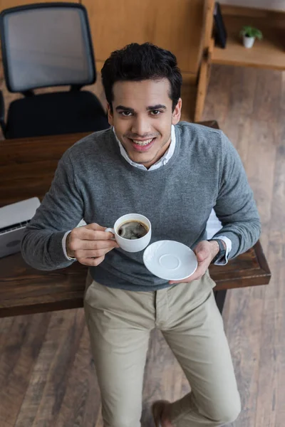 High angle view of businessman with saucer and cup of coffee smiling and looking at camera in office — Stock Photo