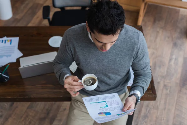 Vista de ángulo alto del hombre de negocios con taza de café mirando el papel cerca de la mesa - foto de stock