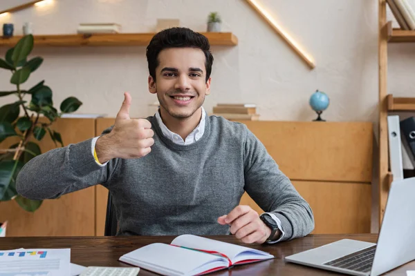 Uomo d'affari guardando la fotocamera, sorridendo e mostrando come segno a tavola in ufficio — Foto stock