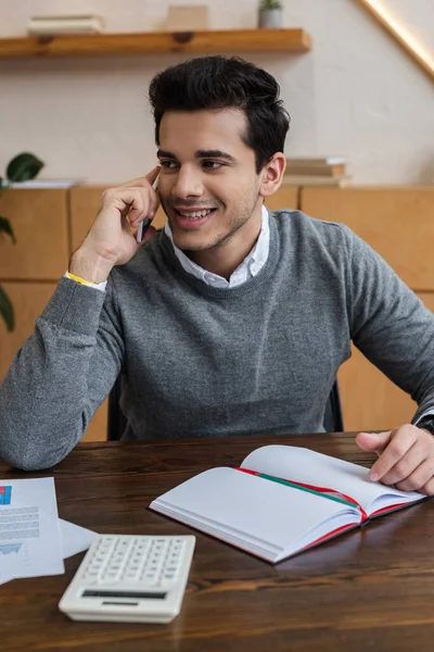 Uomo d'affari che sorride, distoglie lo sguardo e parla su smartphone a tavola in ufficio — Foto stock