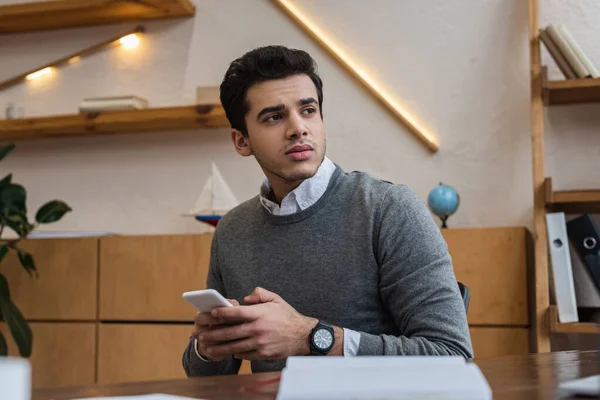 Enfoque selectivo del hombre de negocios reflexivo charlando en el teléfono inteligente y mirando a la mesa en la oficina - foto de stock