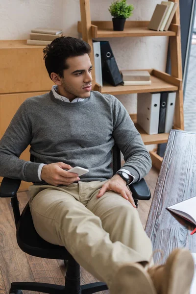 Selective focus of thoughtful businessman with smartphone put legs on table in office — Stock Photo