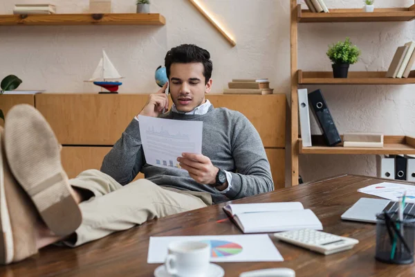 Selective focus of businessman with legs on table looking at paper and talking on smartphone — Stock Photo