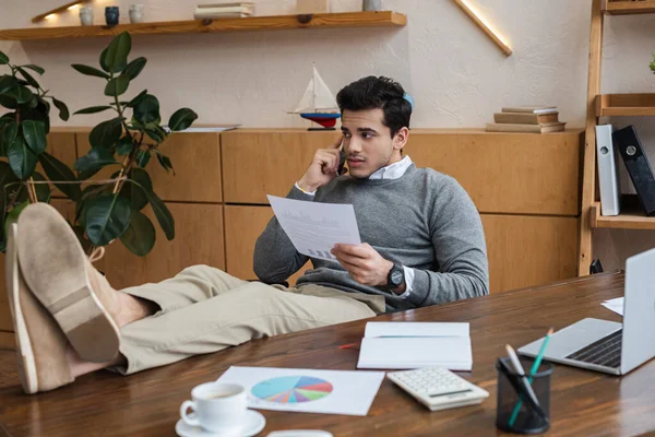 Geschäftsmann mit Papier legt Beine auf den Tisch und spricht im Büro mit Smartphone — Stockfoto