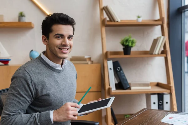 Empresario con tableta digital y lápiz sonriendo y mirando a la cámara en la mesa en la oficina - foto de stock