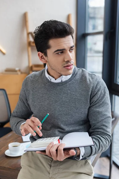 Pensativo hombre de negocios con cuaderno, lápiz y calculadora mirando hacia otro lado en la oficina - foto de stock