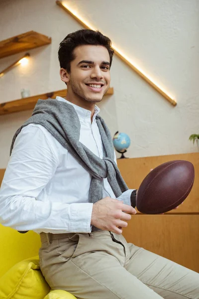 Uomo d'affari con palla da rugby sorridente e guardando la fotocamera sul divano in ufficio — Foto stock