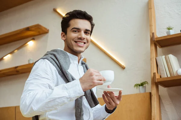 Vista de bajo ángulo del hombre de negocios con platillo y taza de café mirando a la cámara y sonriendo - foto de stock