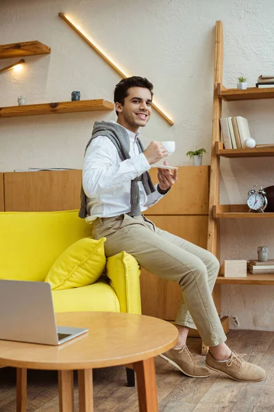 Hombre de negocios con taza de café sonriendo y mirando a la cámara en el sofá cerca de la mesa de café con el ordenador portátil en la oficina - foto de stock