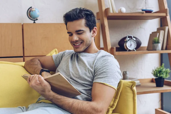 Homem lendo livro e sorrindo no sofá na sala de estar — Fotografia de Stock