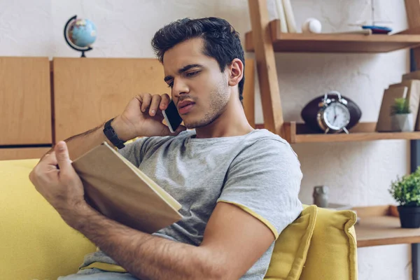 Чоловік з книгою розмовляє на смартфоні на дивані у вітальні — стокове фото