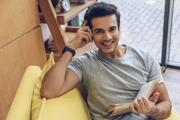 Vista de alto ángulo del hombre con libro sonriendo y mirando a la cámara en el sofá - foto de stock