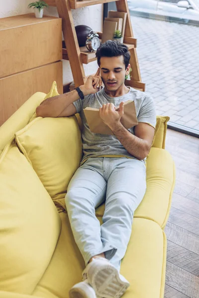 High angle view of man with book talking on smartphone and lying on sofa in living room — Stock Photo