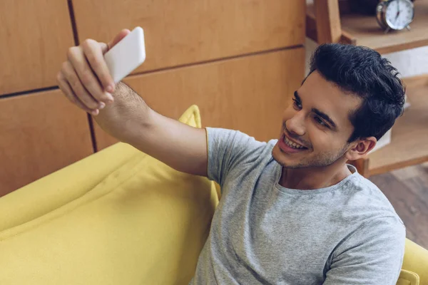 Hombre sonriendo y tomando selfie con teléfono inteligente en el sofá - foto de stock