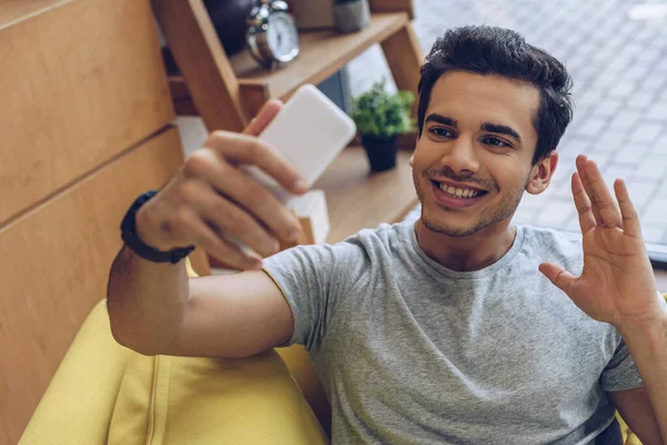 Enfoque selectivo del hombre saludando la mano, sonriendo y tomando selfie con teléfono inteligente en el sofá - foto de stock
